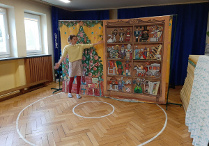 Zdjęcie przedstawia aktorkę przedstawienia na tle choinki i regału z zabawkami.