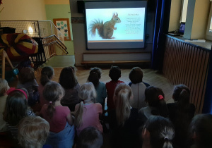 Zdjęcie przedstawia dzieci oglądające prezentację multimedialną. Na slajdzie jest wiewiórka i krótki opis o niej.