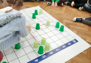 Zdjęcie przedstawia grupę dzieci, które układają zielone kubeczki na odpowiednich polach maty do kodowania.