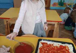 Zdjęcie przedstawia dziewczynkę, która nakłada sos serowy na pizzę.