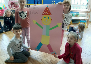 Zdjęcie przedstawia grupę dzieci prezentujących pracę - postać wykonaną z figur geometrycznych.