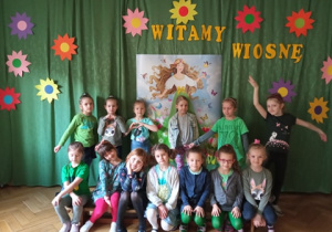 Zdjęcie przedstawia grupę dzieci ubranych na zielono na tle wiosennej dekoracji.