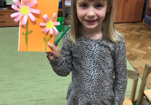 Dziewczynka pokazuje swoją laurkę różowy kwiat na pomarańczowym tle.