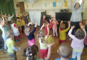 Na zdjęciu grupa dzieci z uniesionymi rękami do góry. Dzieci naśladują ruchy nauczyciela.