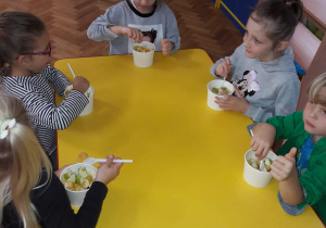 Dzieci jedzą przy stoliku sałatki owocowe w papierowych miseczkach.